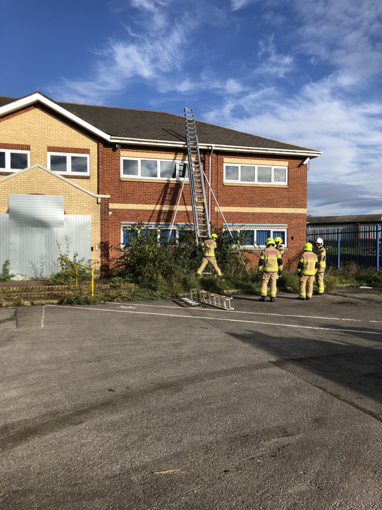 5 fire men stood at the bottom of a ladder which is positioned against the side of a building being prepared for demolition