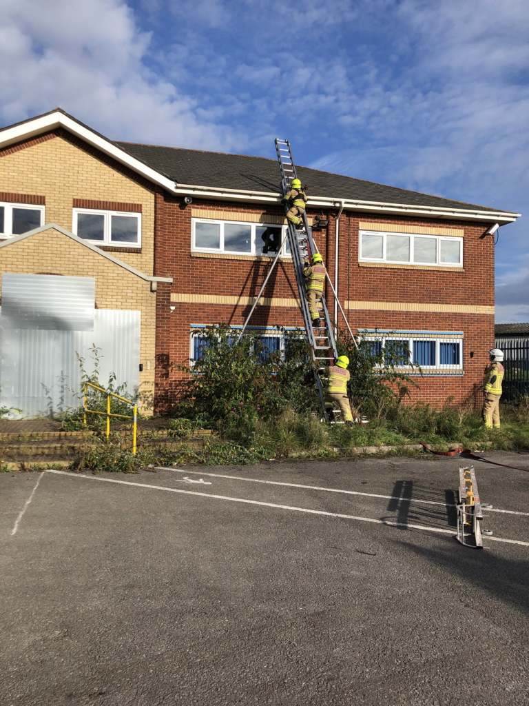 2 firemen have climbed up the ladder and 1 fireman is stood at the bottom of a ladder which is positioned against the side of a building being prepared for demolition